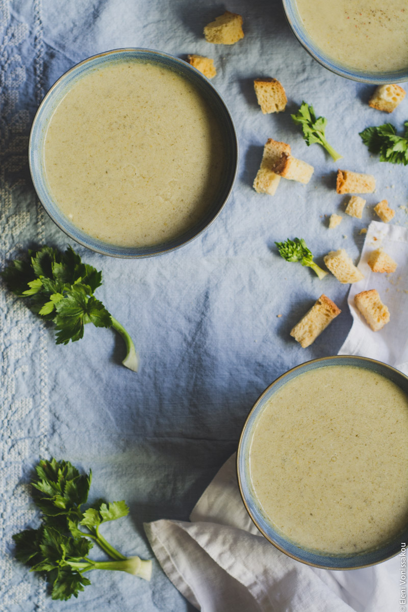 Slow Cooker Cream of Celery Soup with Mascarpone www.thefoodiecorner.gr Photo description: An overhead view of two bowls of soup, sitting on a light blue linen cloth. One bowl also has a white linen napkin underneath it. Scattered around the bowls are small celery leaves and croutons. A third bowl is barely visible in the top right corner.