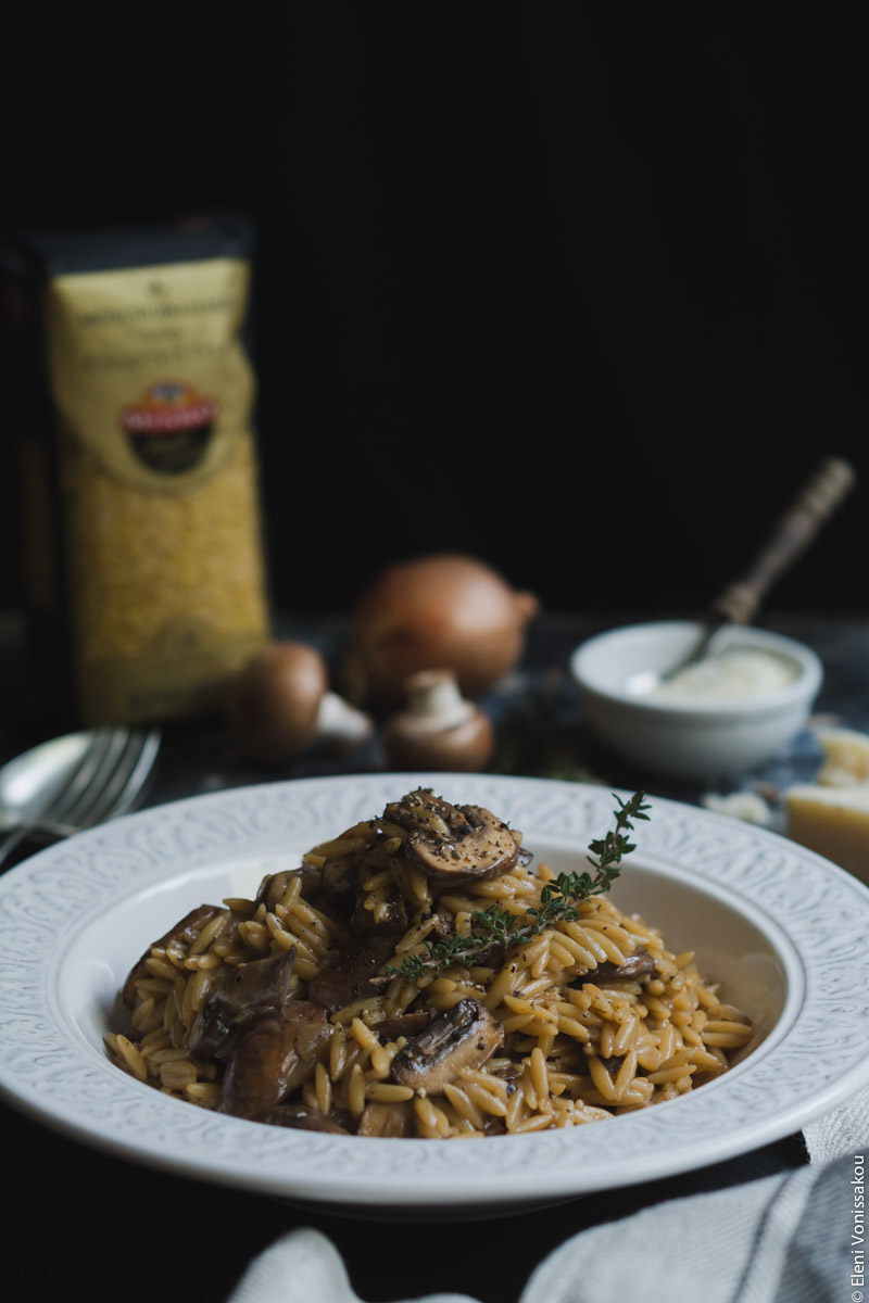 Orzotto with Mushrooms, Balsamic Cream and Parmesan www.thefoodiecorner.gr Photo description: A side view of the orzotto, on a white plate with a sprig of thyme on top. In the background to the left barely visible, an upright packet of orzo, an onion and two mushrooms, and a bowl of grated parmesan to the right. The backdrop is black.