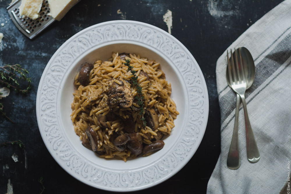 Orzotto with Mushrooms, Balsamic Cream and Parmesan www.thefoodiecorner.gr Photo description: A closer view of the plate of orzotto. A slice of mushroom visible on top of the pasta, with a sprig of thyme. To the right of the plate a fork and spoon lying on a bunched up linen tea towel. Barely visible to left above the plate, a broken piece of parmesan. All sitting on a dark surface.