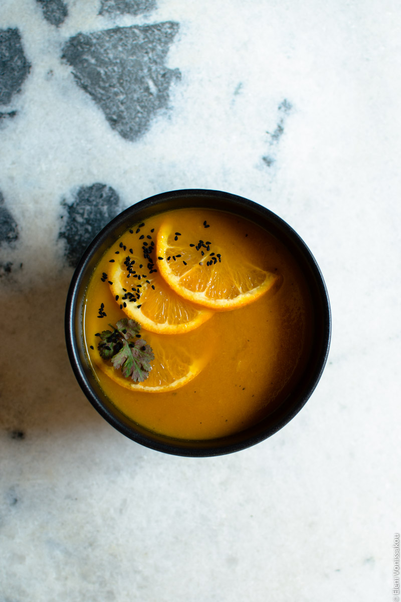 Simple Slow Cooker Carrot and Orange Soup with Turmeric and Cumin www.thefoodiecorner.gr Photo description: A black bowl with carrot and orange soup, sitting on a marble surface, in the centre of the photo. On the soup, a garnish of three slices of orange, one coriander leaf and some nigella seeds. The marble surface has some grey spots towards the top left corner, the rest is white.