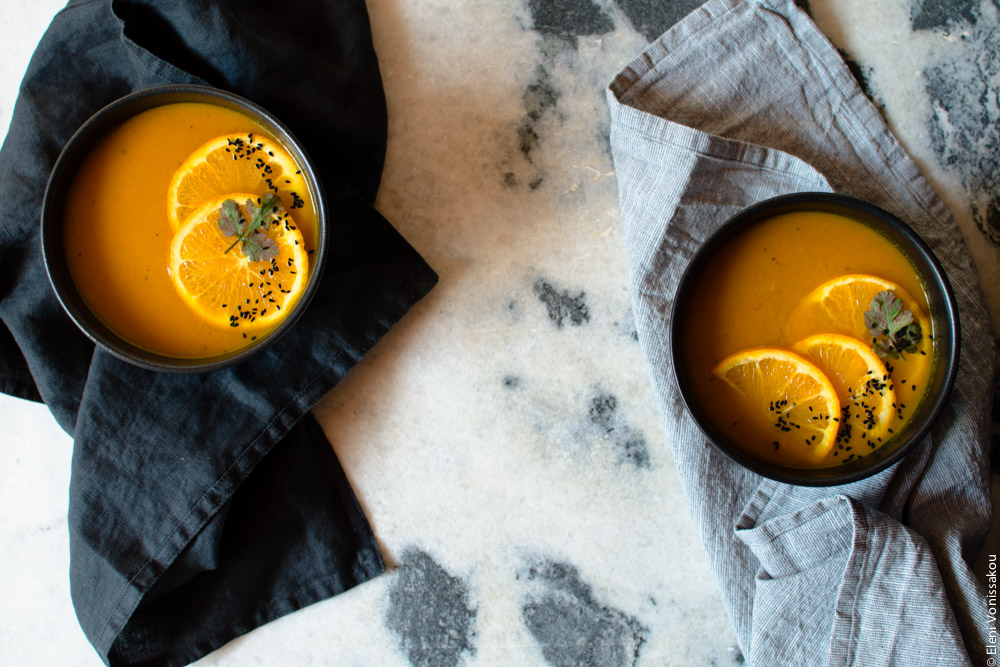 Simple Slow Cooker Carrot and Orange Soup with Turmeric and Cumin www.thefoodiecorner.gr Photo description: Two black bowls of carrot orange soup, at each end of the photo (landscape layout). One is sitting on a bunched up black linen napkin, the other on a grey dish cloth. The grey and white marble surface is visible in the middle. Garnishes as in other pics.