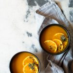 Simple Slow Cooker Carrot and Orange Soup with Turmeric and Cumin www.thefoodiecorner.gr Photo description: Two black bowls with carrot and orange soup, on a grey and white marble surface. One bowl is sitting on a grey linen dish cloth. Both bowls have a garnish of orange slices, a coriander leaf and some nigella seeds.