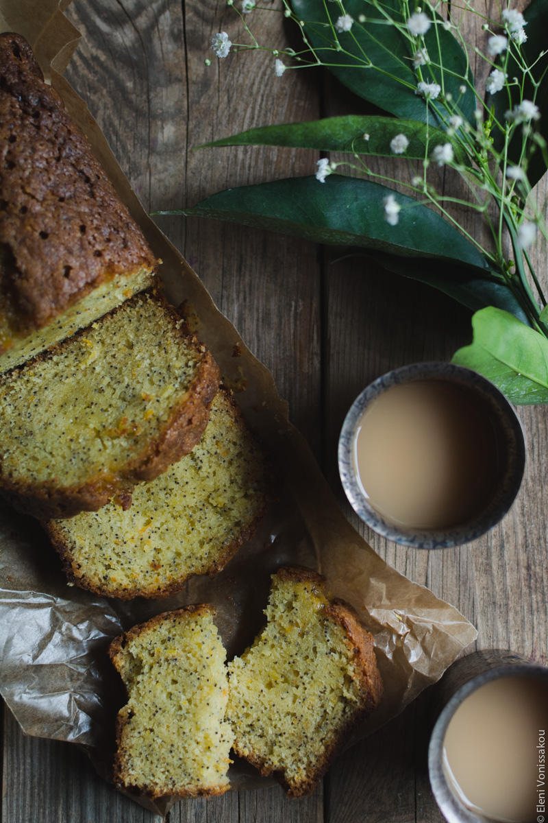 Sticky Orange Poppy Seed Olive Oil Loaf Cake www.thefoodiecorner.gr Photo description: The cake lying diagonally from top left to the centre of the photo. The main part is only partly visible, and there are two whole slices lying in front. Further down is one slice broken in two. To the right of the cake are two metal cups with tea in them. In the top right corner some orange leaves and small white flowers.