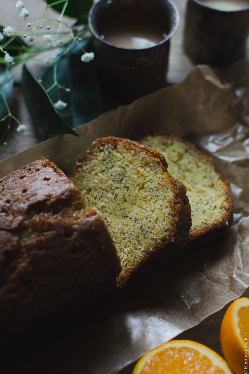 Sticky Orange Poppy Seed Olive Oil Loaf Cake www.thefoodiecorner.gr Photo description: A close view of the top half of the cake, with two cut slices lying in front of it. Cake is on brown baking paper. To the top of the photo two metalic cups with tea and a couple of leaves from an orange tree. To the bottom of the photo barely visible, two halves of an orange.