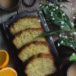 Sticky Orange Poppy Seed Olive Oil Loaf Cake www.thefoodiecorner.gr Photo description: Slices of cake lying on a silver rectangular tray, sitting on a wooden surface. To the right, a branch from an orange tree and some small white flowers. In the top right corner a ball of string and a pair of black scissors. In the top left corner a metal cup with tea, and to the left of the tray an orange cut in half. Another cup of tea is barely visible in the bottom right corner.