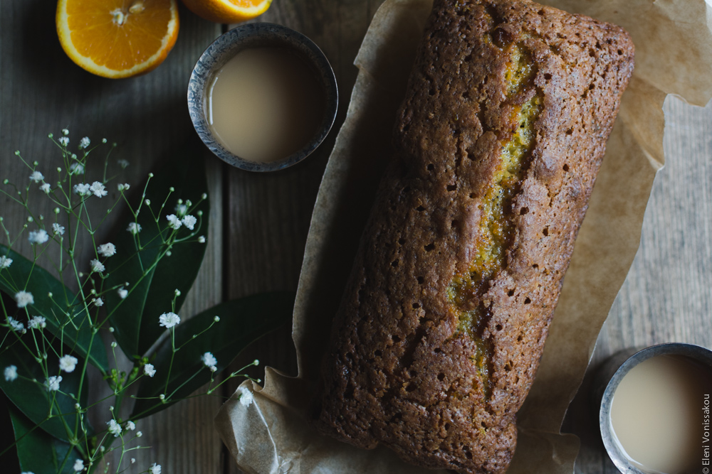 Sticky Orange Poppy Seed Olive Oil Loaf Cake www.thefoodiecorner.gr Photo description: The cake before it was sliced, sitting on brown baking paper on a wooden surface. To its left and right, two cups of tea. To the far left of the photo orange leaves and some small white flowers. Barely visible in the top left corner two halves of an orange.