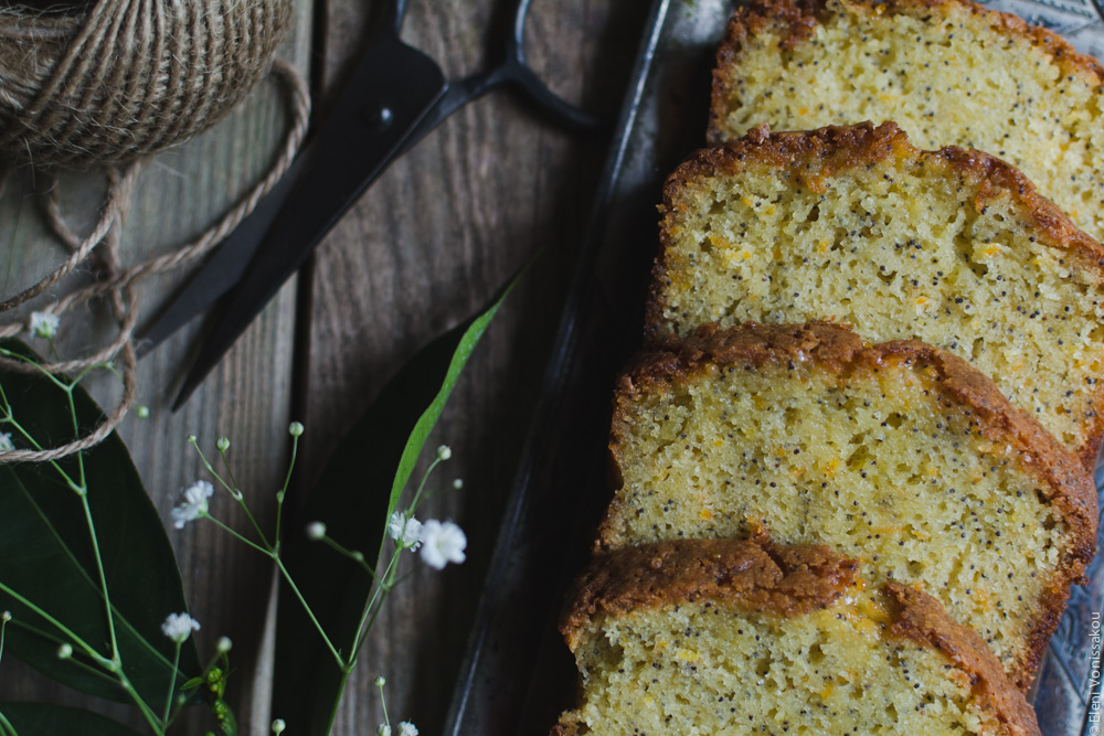 Sticky Orange Poppy Seed Olive Oil Loaf Cake www.thefoodiecorner.gr Photo description: A close up of the slices of cake on the silver tray. The cake is brown around the egdes and moist and yellowish on the inside. Small flecks of black poppy seeds are visible. To the left of the photo some orange leaves, the ball of string and the black scissors, all half visible.