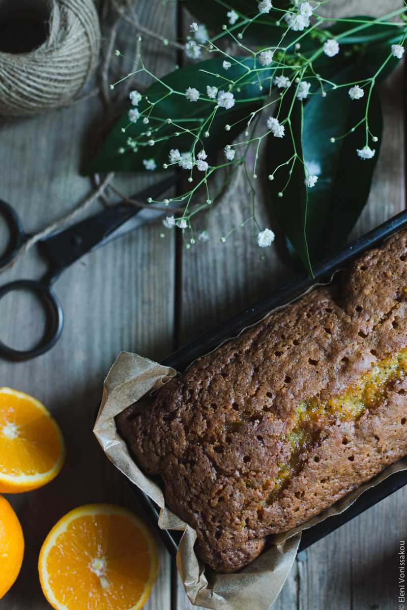 Sticky Orange Poppy Seed Olive Oil Loaf Cake www.thefoodiecorner.gr Photo description: The cake, half visible, still in the loaf pan with brown baking paper showing around the edges. To the top of the photo the leaves and flowers, next to them the string and scissors. In the bottom left corner the orange halves, and a whole orange barely visible.