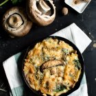 Pasta Bake with Spinach and Portobello Mushrooms in Bechamel Sauce www.thefoodiecorner.gr Photo description: A black cast iron skillet with a golden crunchy topped pasta bake, sitting on a folded linen tea towel. To the top left are two Portobello mushrooms lying stalk-up and some spinach leaves, to the centre are some raw penne pasta pieces, and to the right is a small porcelain grater with some nutmeg on it. All on a black rough and slightly battered surface.