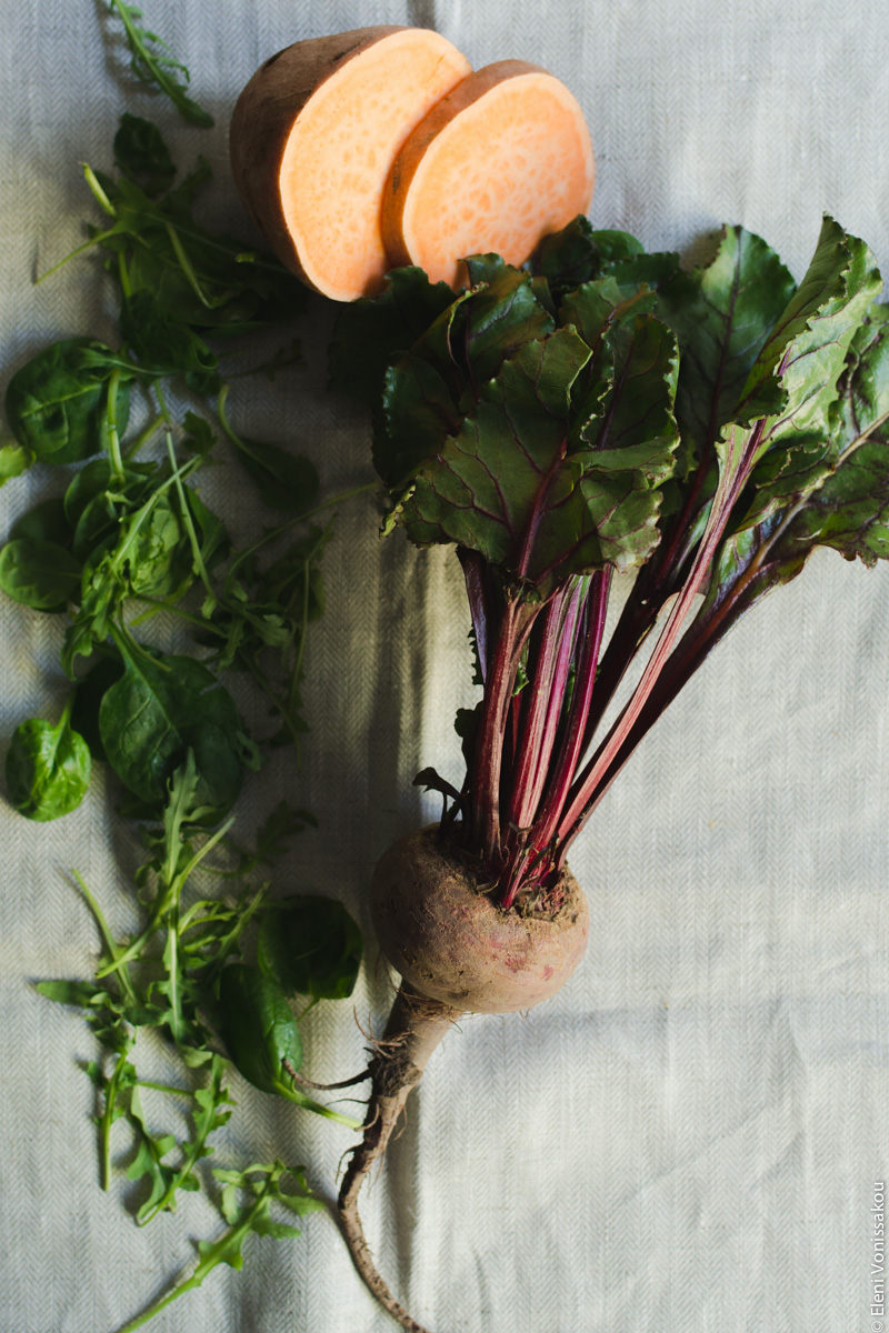 Roast Beetroot and Sweet Potato Buddha Bowl with Spicy Tahini Honey Dressing www.thefoodiecorner.gr  Photo description: Ingredient shot with a whole beetroot (with leaves) lying on the linen material. To the top half a sweet potato and a cut slice, and to the left of the beetroot, some scattered greens (spinach and rocket).