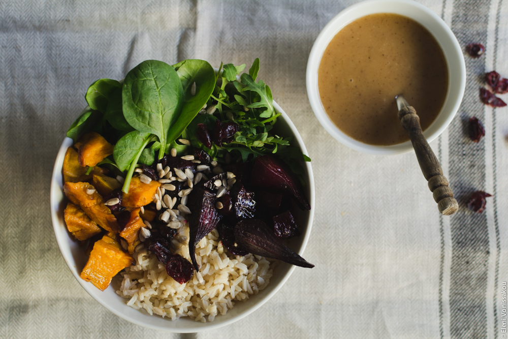 Roast Beetroot and Sweet Potato Buddha Bowl with Spicy Tahini Honey Dressing www.thefoodiecorner.gr  Photo description: A close up of the one bowl and its contents. To the right the small bowl of dressing with a spoon in it. The spoon has a wooden handle. Some scattered cranberries are also visible to the right of the dressing, almost out of shot.