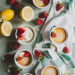 Easy Baked Lemon Puddings www.thefoodiecorner.gr Photo description: Top view of five lemon puddings in white ramekins, spread out on a light green wooden surface. Two of them are sitting on a scrunched up linen tea towel. Scattered here and there between the ramekins are whole strawberries and camomile flowers. At the top of the photo is a small sieve with icing sugar, and to the left are one whole and one half lemon. Near that, a bowl of mascarpone cheese with some strawberries on top. The puddings have sprinkled icing sugar and some strawberries on them.