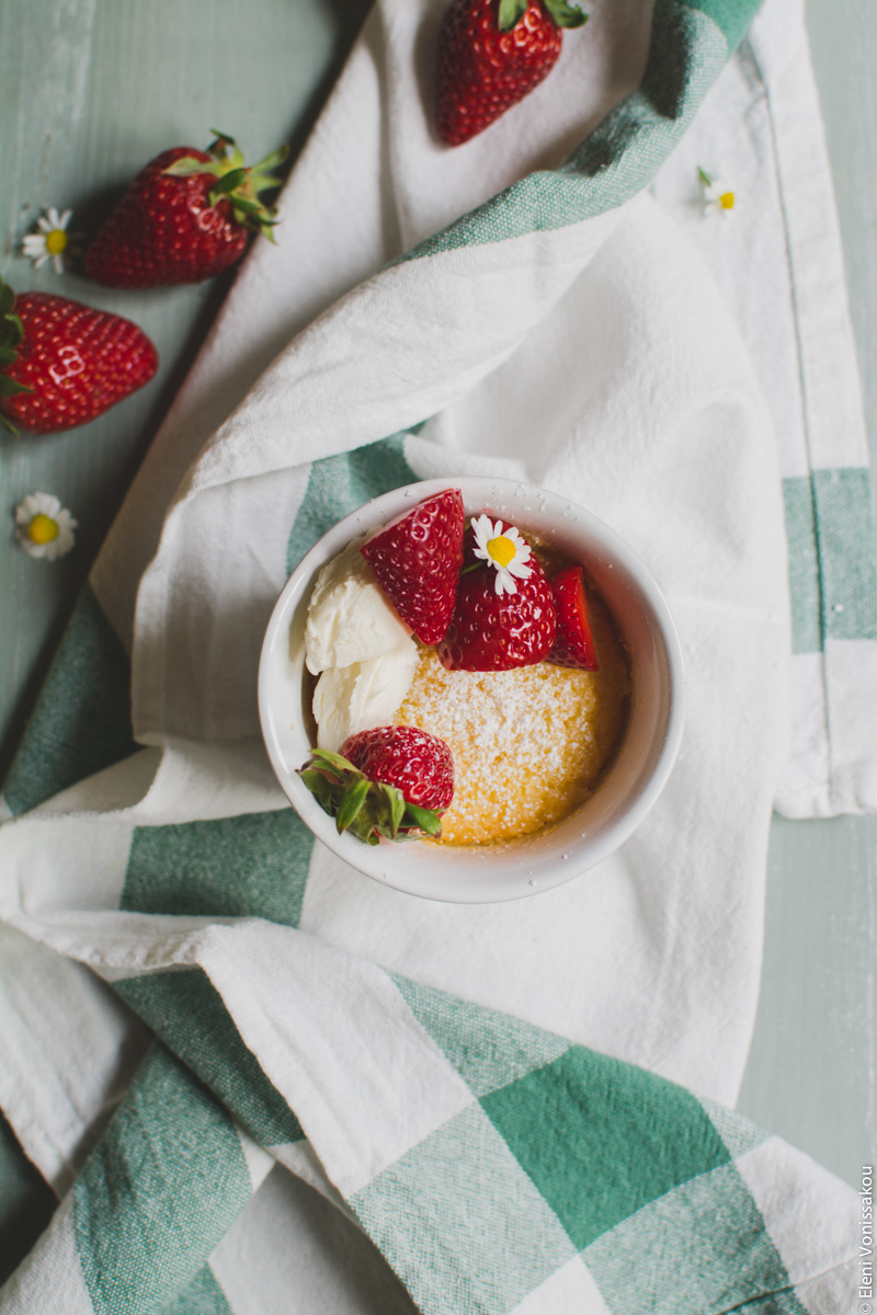 Easy Baked Lemon Puddings www.thefoodiecorner.gr Photo description: One lemon pudding sitting on a scrunched up linen tea towel. The top is sprinkled with icing sugar and there are some strawberries, mascarpone cheese and a small camomile flower decorating the pudding. To the top left of the photo are some scattered strawberries on the wooden surface.