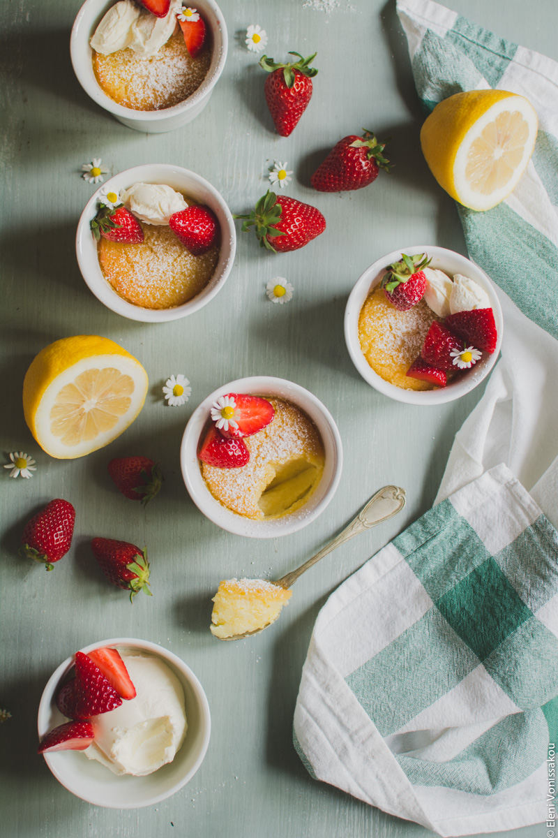 Easy Baked Lemon Puddings www.thefoodiecorner.gr Photo description: The decorated puddings sitting on the wooden surface, with scattered strawberries and two lemon halves around them. A spoon is lying next to one of the ramekins with a dollop of the pudding on it. 