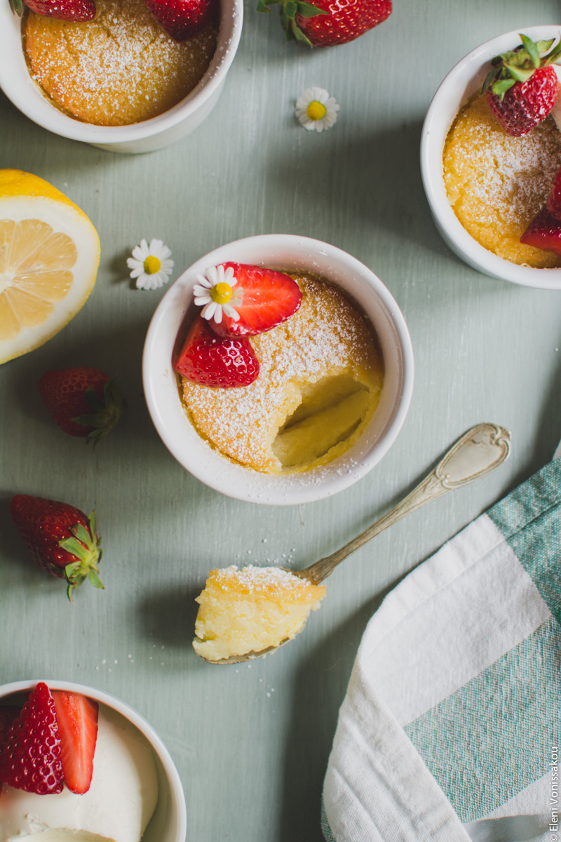 Easy Baked Lemon Puddings www.thefoodiecorner.gr Photo description: A closer view of the one ramekin with a spoonful of creamy yellow pudding lying beside it. The pudding is decorated with two pieces of strawberry and a little camomile flower.