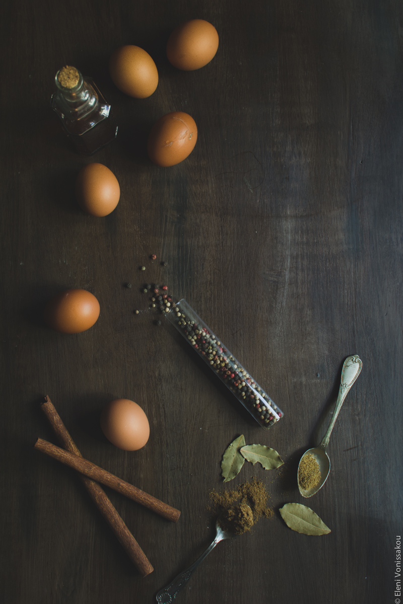 Beetroot Pickled Eggs www.thefoodiecorner.gr Photo description: An overhead view of ingredients spread out on a dark wooden surface. A bottle of vinegar, eggs, cinnamon sticks, bay leaves, spoons with spices on them and a glass vial with colourful peppercorns.