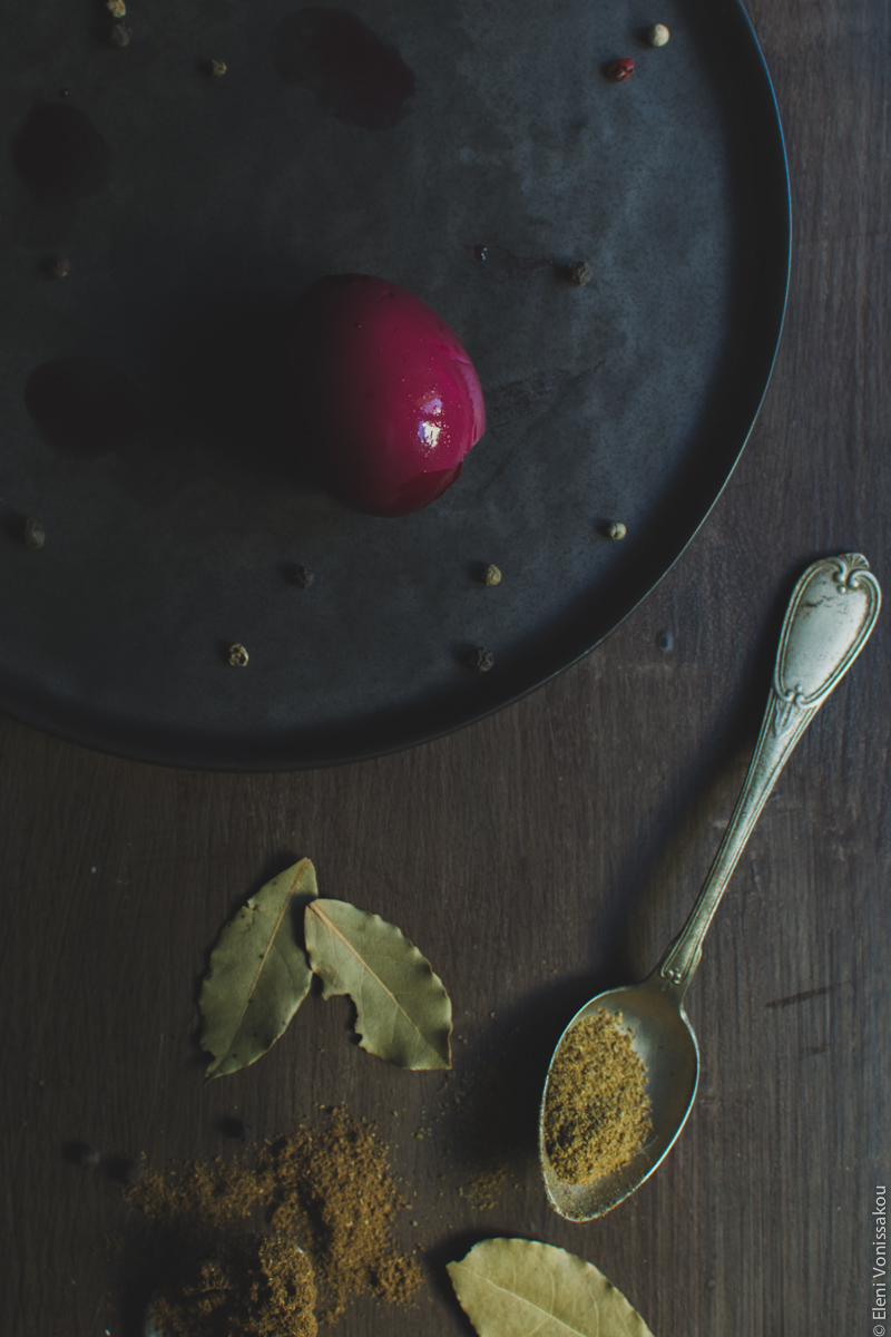 Beetroot Pickled Eggs www.thefoodiecorner.gr Photo description: The black plate with one whole pickled egg on it. The egg has turned a dark purple colour from the beetroot juice. Towards the bottom of the photo are some bay leaves and some spices lying in spoons on the wooden surface.