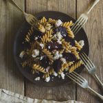 Roast Beetroot and Feta Pasta Salad with Greek Yoghurt and Cumin Dressing www.thefoodiecorner.gr Photo description: a brown plate of fusilli pasta salad on a weather beaten wooden surface. At the bottom of the photo, a linen napkin just visible. Around the plate, four forks are propped face down against the edge.