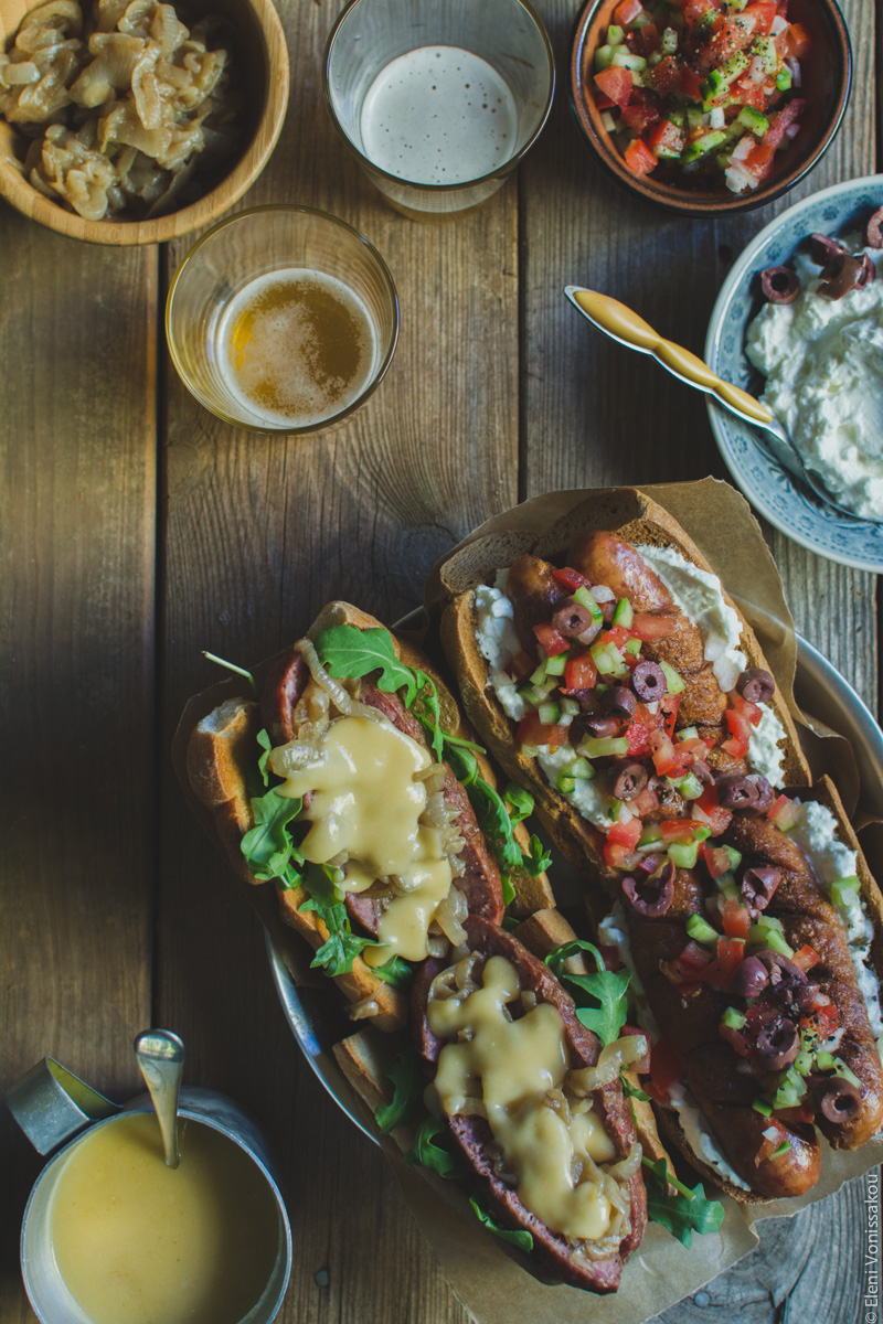 Two Ways with Greek Sausage Hot Dogs www.thefoodiecorner.gr - Photo description: A top view of the plate of hot dogs, this time with four (2 of each flavour) arranged on the baking paper. The plate is positioned to the bottom right, and the condiments and beers are in the top of the photo. Only the cheese sauce is in the bottom left corner.