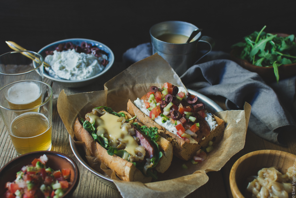 Two Ways with Greek Sausage Hot Dogs www.thefoodiecorner.gr - Photo description: A ¾ view of the plate of hot dogs. The bowls of condiments are arranged around it together with a couple of glasses of beer. Everything is on an old wooden surface set against a dark backdrop.