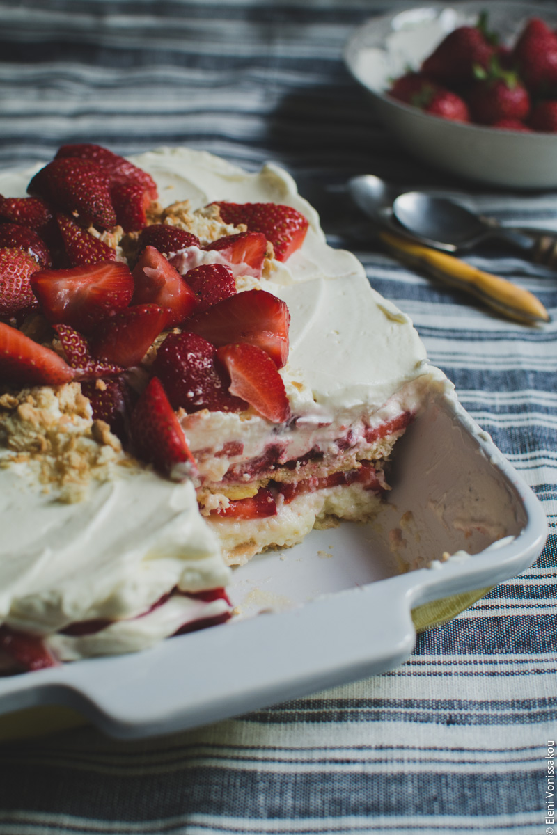 Strawberries and Cream Fridge (Icebox) Cake www.thefoodiecorner.gr Photo description: Close up of the baking dish from a ¾ angle, with a piece of cake missing from the front corner. The layers of the cake are visible from the side. In the background to the right the bowl of strawberries.