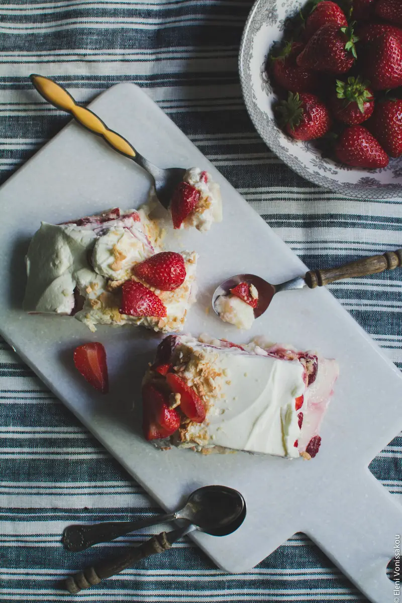 Strawberries and Cream Fridge (Icebox) Cake www.thefoodiecorner.gr Photo description: Two pieces of cake on a white marble cutting board. The pieces are sloppy and some of the cream is spilling over. Two spoons with spoonfulls of cake are lying next to the pieces. To the top right the bowl of strawberries.