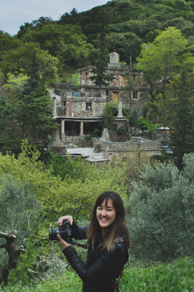 Milia Workshop 2017 – A long overdue recap. Part 1. www.thefoodiecorner.gr - Photo description: A young lady standing sideways with her face turned to the camera. She is holding her own camera aimed in front of her. In the background, the stone buildings of Milia among green trees and bushes.