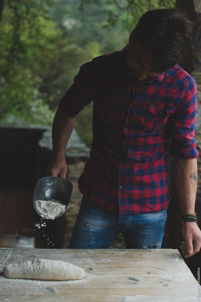 Miliaworkshop2017 www.thefoodiecorner.gr Photo description: Front view of a young man in jeans and a checked shirt, standing behind a wooden table, sprinkling flour from a metal scoop over some dough shaped into a loaf.