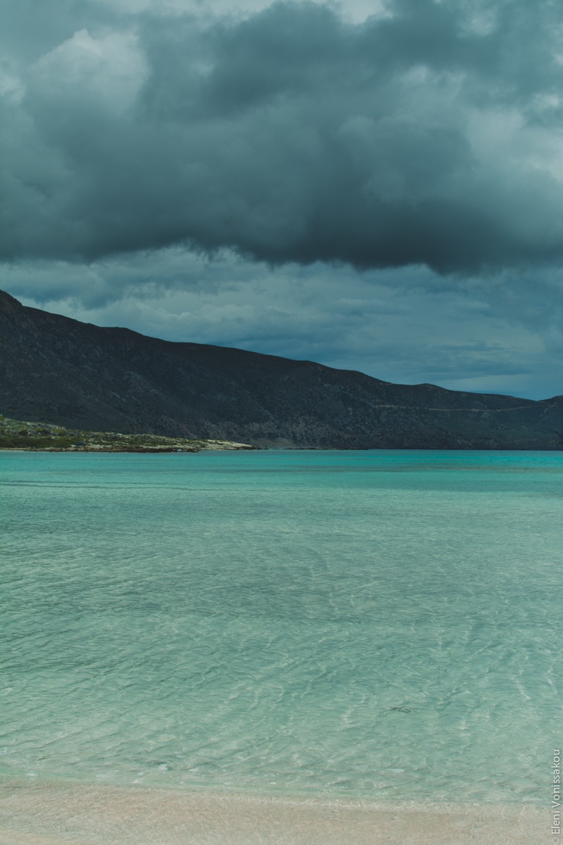 Miliaworkshop2017 www.thefoodiecorner.gr Photo description: the turquoise shallow waters of the sea, from the light coloured sand within, and a dark sky above with low heavy clouds. A mountain side is visible in the distance.