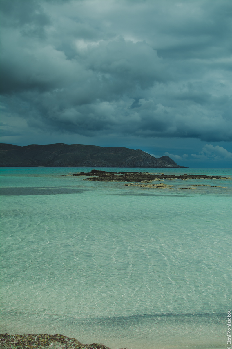 Milia Workshop 2017 – A long overdue recap. Part 1. www.thefoodiecorner.gr - Photo description: Crystal clear shallow sea with white sand visible. To the middle of the photo some rocks protruding from the turquoise waters, and a mountain in the distance. The sky is dark with heavy gray clouds.