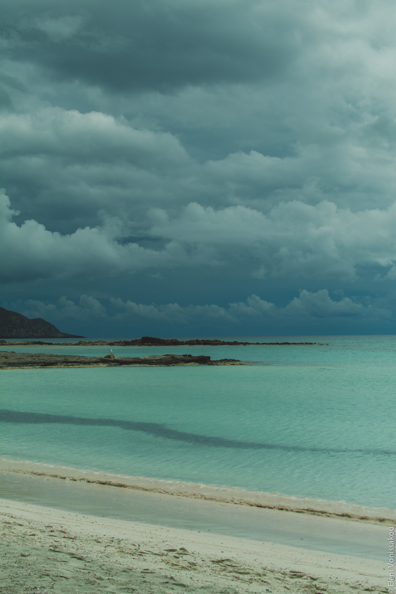 Miliaworkshop2017 www.thefoodiecorner.gr Photo description: The beach and sea, with light coloured sand, turquoise waters and a dramatic sky with dark heavy clouds in the distance.