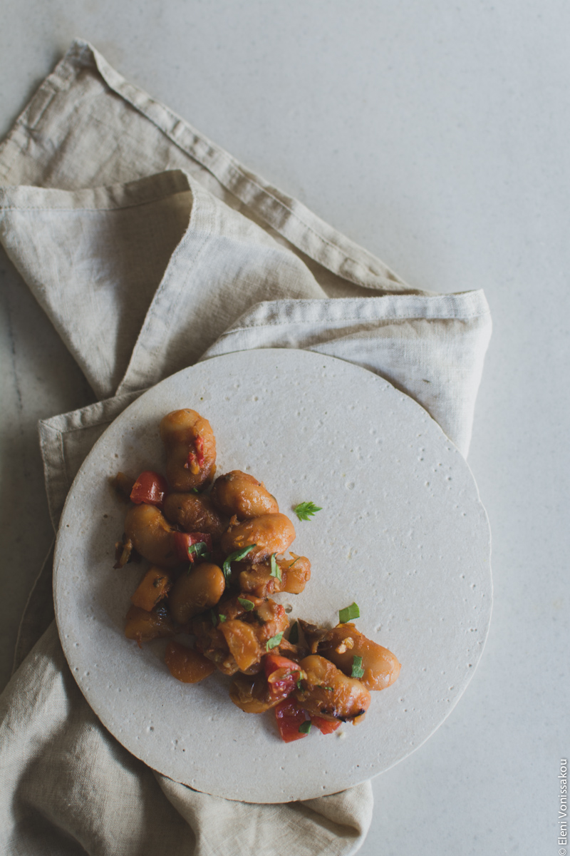 Miliaworkshop2017 www.thefoodiecorner.gr Photo description: a cement plate with some giant beans cooked in tomato sauce, laid to one side of it. The plate is sitting on a light coloured linen napkin, and everything is on a marble surface.