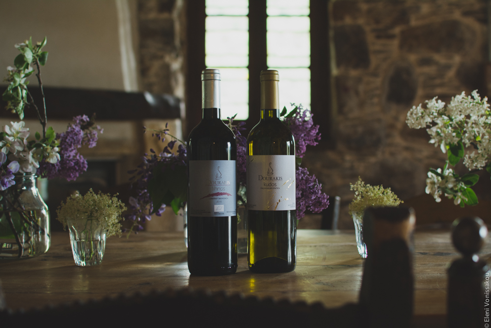 Miliaworkshop2017 www.thefoodiecorner.gr Photo description: two bottles of Dourakis wine on a big wooden table, among several vases with flowers. In the background a stone wall and a window with a wooden pane.