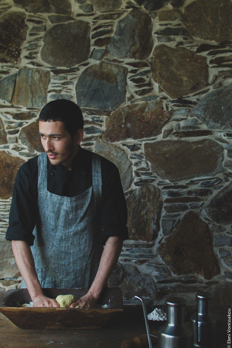 Miliaworkshop2017 www.thefoodiecorner.gr Photo description: A young man standing behind a wooden table, pressing down on some dough in a wooden basin. He is wearing a linen apron and looking to his right. Behind him a stone wall.