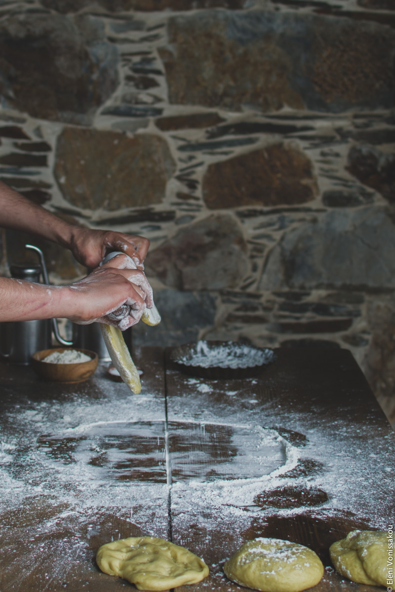 Miliaworkshop2017 www.thefoodiecorner.gr photo description: A side view of a pair of floured hands holding a rolling pin. There is some partly rolled out dough hanging from the rolling pin. The table beneath is covered in flour and there are a few rounds of dough on it waiting to be rolled out. In the background a stone wall.