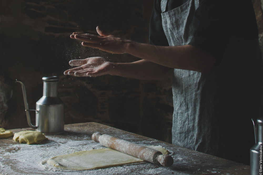 Miliaworkshop2017 www.thefoodiecorner.gr Photo description: A table with some rolled out dough and a rolling pin on it, and an open pair of hands mid air looking as if they have just been clapped together. Particles of flour are visible flying around in between the hands.