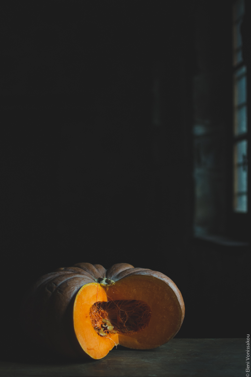 Milia Workshop 2017 – A long overdue recap. Part 1. www.thefoodiecorner.gr - Photo description: A large pumpkin with a quarter of it cut out, revealing the seeds and fibres in the centre. The pumpkin is on a wooden surface, lit from the right from a window that is barely visible. The light is brightening the vivid orange colour of one of the cut sides. The background is in darkness.