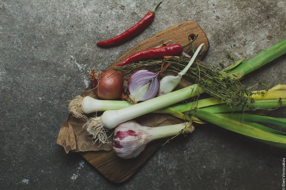 Miliaworkshop2017 www.thefoodiecorner.gr Photo description: a wooden chopping board with onions, leeks, peppers and herbs on it, all on a cement surface.