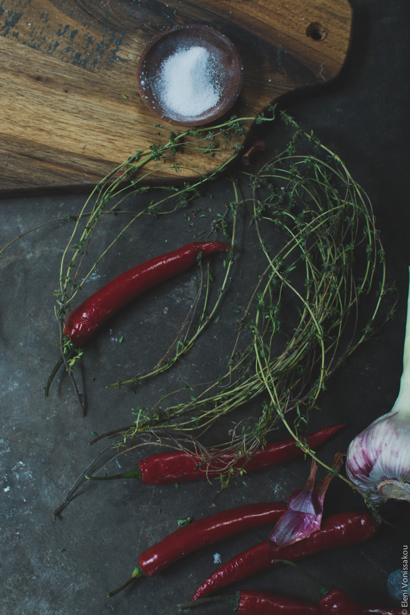 Miliaworkshop2017 www.thefoodiecorner.gr Photo description: at the top of the photo a Tzoumani Bros chopping board half visible, with a tiny bowl of salt on it. Below some fresh thyme, red chilli peppers, and some fresh garlic barely visible, all on a rough cement surface.