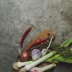 Milia Workshop 2017 – A long overdue recap. Part 1. www.thefoodiecorner.gr - Photo description: A chopping board with some fresh garlic, onions, leeks, thyme and red pepper on a piece of grease proof paper. The board is towards the bottom of the photo. A single red pepper lies next to the board. Everything is on a rough gray surface. To the top of the photo only the gray surface is visible.