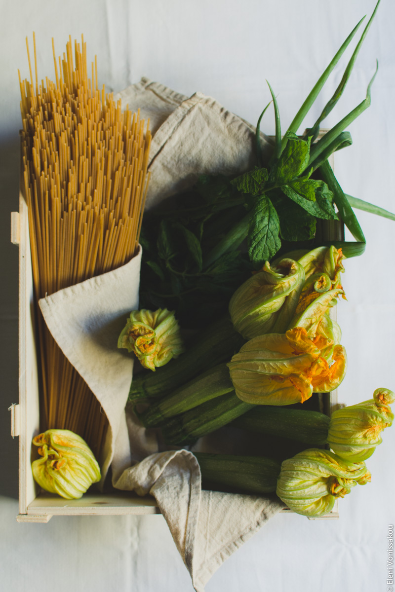 Whole Wheat Courgette Pasta with Courgette Flowers www.thefoodiecorner.gr Photo description: An open wooden box lined with a linen tea towel, containing whole courgettes with their flowers, propped up against the right side. There is also a bunch of spring onions and some sprigs of spearmint in the box next to the courgettes. Lying in the box on its left side, is some uncooked whole wheat spaghetti.