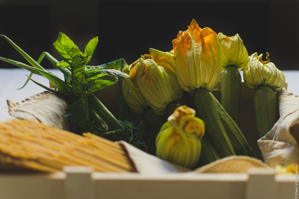 Whole Wheat Courgette Pasta with Courgette Flowers www.thefoodiecorner.gr Photo description: a wooden box lined with a linen teatowel, containing courgettes with their flowers, spring onions and spearmint, and also some uncooked wholewheat spaghetti.  The box is on a cotton tablecloth and the light is coming in from behind it, lighting up the courgette flowers nicely.