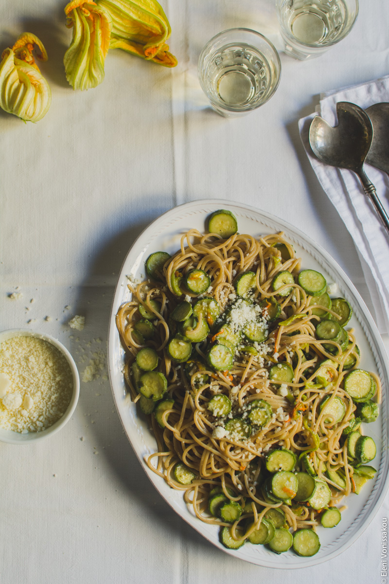 Whole Wheat Courgette Pasta with Courgette Flowers www.thefoodiecorner.gr Photo description: the platter of spaghetti with a couple of glasses of wine visible at the top of the photo, next to some courgette flowers. To the left of the platter is a small bowl of cheese. There is also cheese on the creased cotton tablecloth. To the right are the serving spoons on a folded linen napkin.