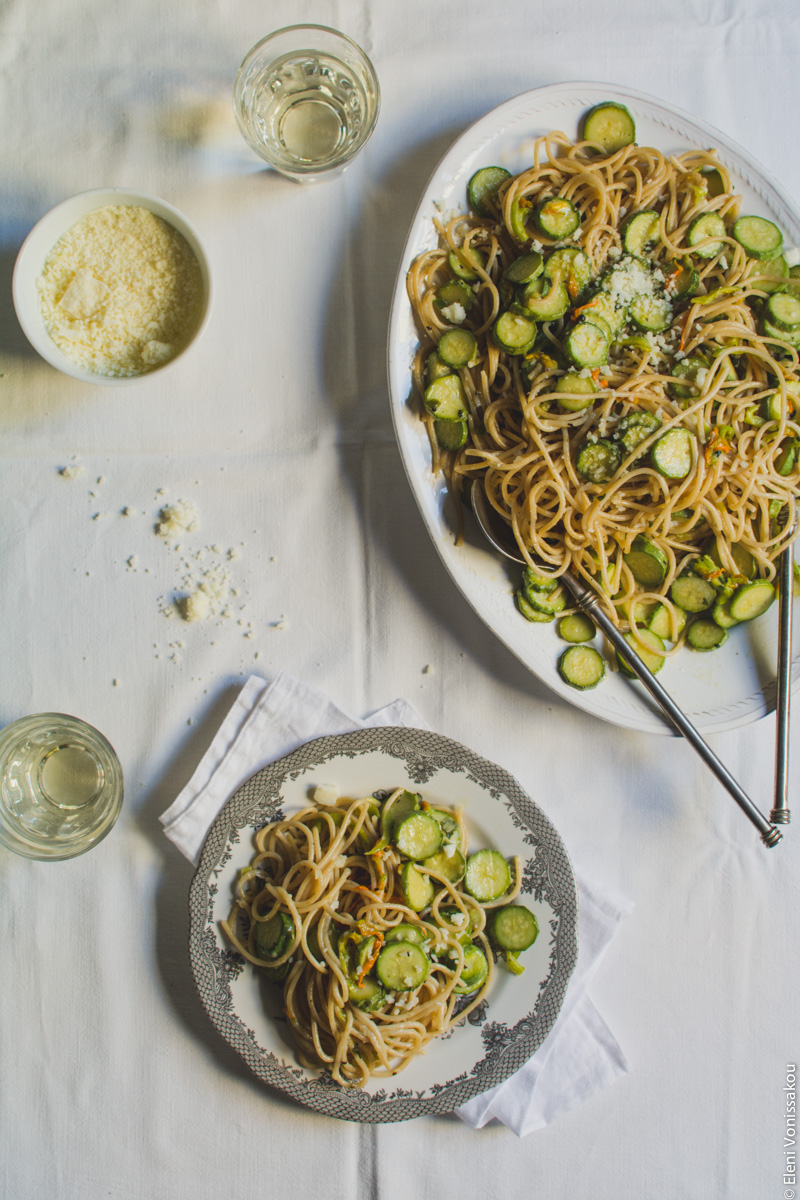 Whole Wheat Courgette Pasta with Courgette Flowers www.thefoodiecorner.gr Photo description: The platter of spaghetti in the top right corner, and a small plate with a pattern around the edge in the bottom centre of the photo. The plate is sitting on a folded linen napking, and has some courgette pasta on it. The serving spoons are lying in the large platter. To the top left of the photo is a glass of wine and a small bowl of grated cheese. There another glass of wine next to the plate on its left.