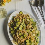 Whole Wheat Courgette Pasta with Courgette Flowers www.thefoodiecorner.gr Photo description: An oval shaped platter with courgette spaghetti on it. The platter is towards the bottom right corner of the photo. To the top right are a set of serving spoons on a linen napkin. In the corner above – barely visible – is a small bowl with grated cheese. In the top left corner are some courgette flowers and two short glasses with white wine. Everything is on a white, slightly creased, cotton tablecloth.