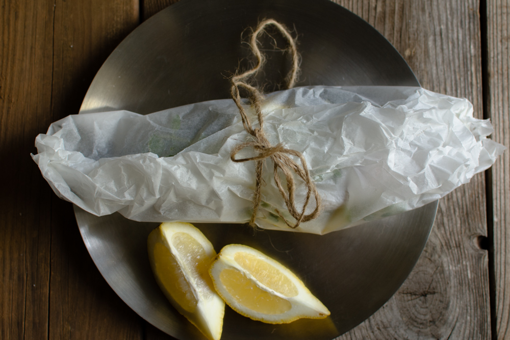 Gilt Head Bream (Dorade) – How to cook a whole fish in the slow cooker www.thefoodiecorner.gr Photo description: the fish (not visible) wrapped in the baking paper. The packet is lying across the metal plate, a piece of string holding it in place. To the bottom of the packet are some lemon wedges.