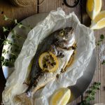 Gilt Head Bream (Dorade) – How to cook a whole fish in the slow cooker www.thefoodiecorner.gr Photo description: A cooked whole fish, lying on baking paper on a metallic plate. On it is a slice of lemon and a couple of sprigs of oregano, both cooked down and wilted. To the top left of the photo a ball of string, to its right a vintage pair of scissors, further to the right a couple of wedges of lemon. In the bottom right corner a small ceramic bowl of salt and a sprig of fresh oregano. Some more lemon wedges and oregano are around the fish to decorate it. Everything is on a wooden surface.