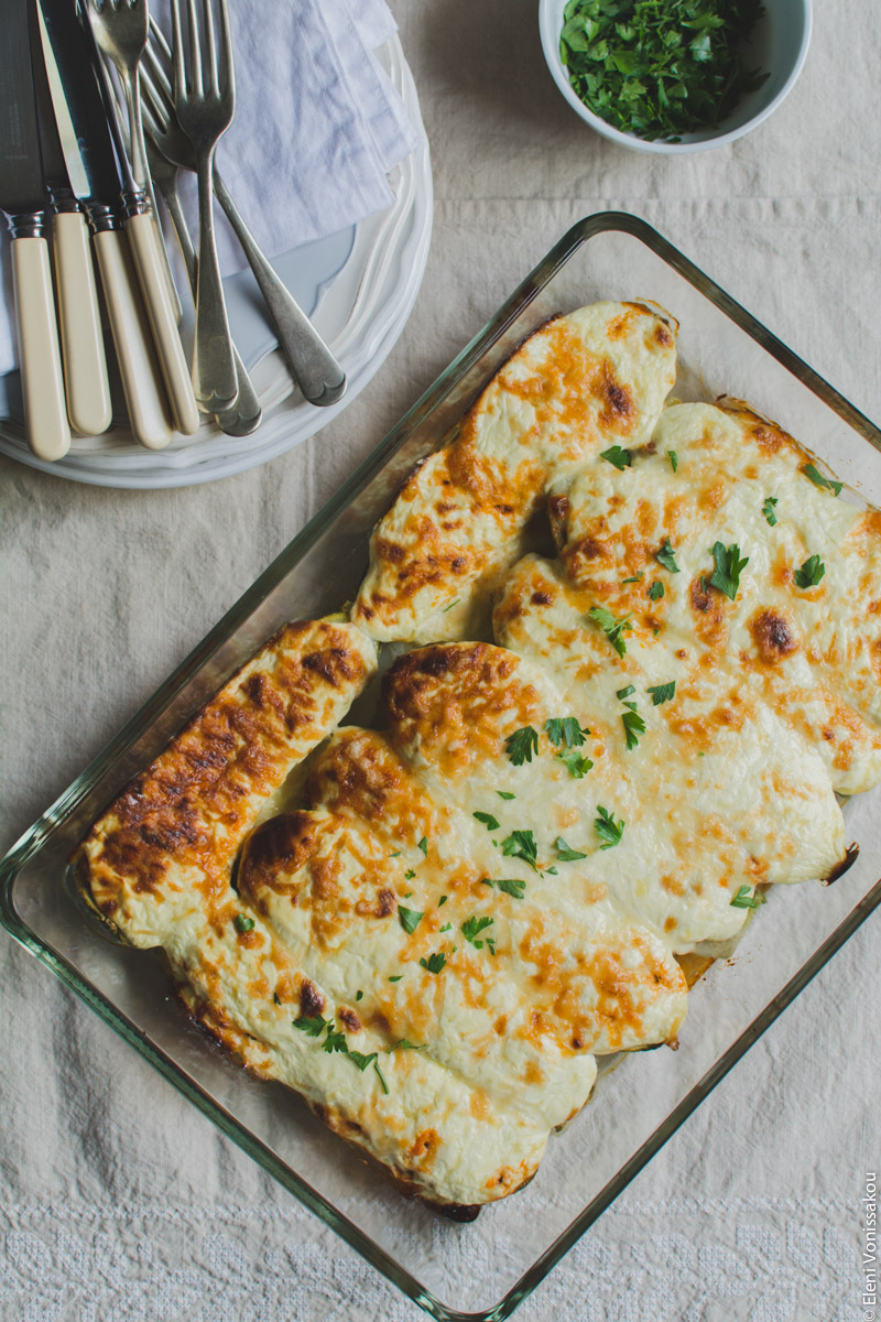 Courgette (Zucchini) Boats with Chicken and Greek Yoghurt “Bechamel” www.thefoodiecorner.gr Photo description: The baking dish with the courgette boats just out of the oven. The top is crispy and golden, with some fresh parsley scattered on it to decorate. To the top left a couple of stacked plates with a napkin on them and some knives and forks. To the top right a bowl of chopped parsley.