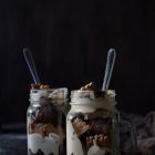 Olive Oil Brownie and No-Churn Coffee Mascarpone Ice Cream Sundae www.thefoodiecorner.gr Photo description: A side view of two jars with brownie ice cream sundaes. Layers of brownie and ice cream are visible as well as broken biscuits within the jars. Spoons are sitting in the jars, with the handles protruding from the top. The sundaes are sitting on a rough dark surface with a dark background behind them.