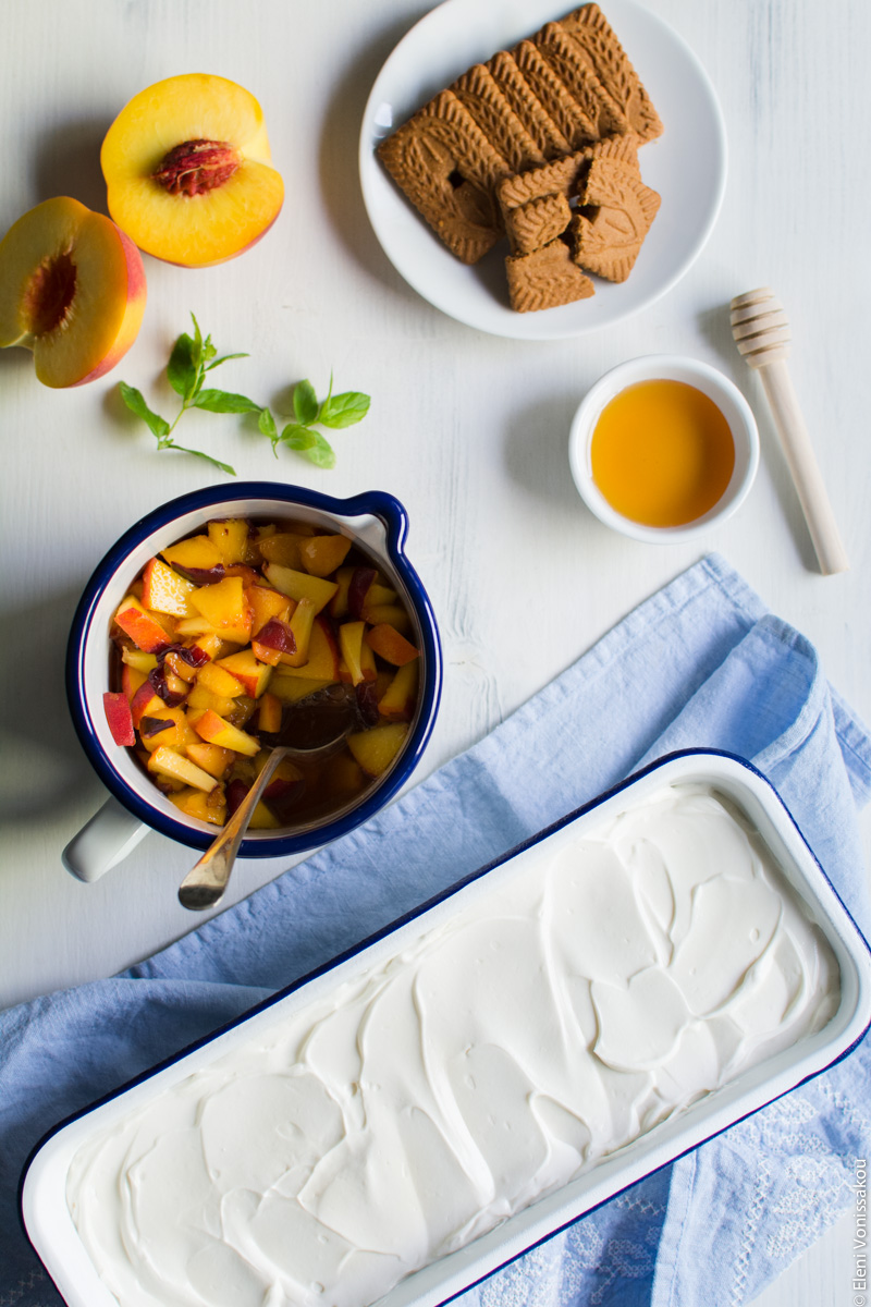 Greek Yoghurt, Mascarpone and Speculoos (Biscoff) Cookie Fridge Cake with Peaches www.thefoodiecorner.gr Photo description: The fridge cake before being decorated, just a white creamy surface in the loaf pan. Further up a jug of chopped peaches, a plate of cookies, a peach cut in half and a small bowl with honey.